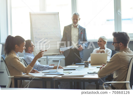 Two diverse office workers talking and smiling while having a meeting with colleagues in the modern office, mature businessman giving a presentation on the background Two diverse office workers talking and smiling while having a meeting with colleagues in the modern office, mature businessman giving a presentation on the background 70868126