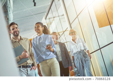 Two young happy diverse male and female coworkers discussing something and smiling while walking with colleagues in the modern office or coworking space Two young happy diverse male and female coworkers discussing something and smiling while walking with colleagues in the modern office or coworking space 70868128