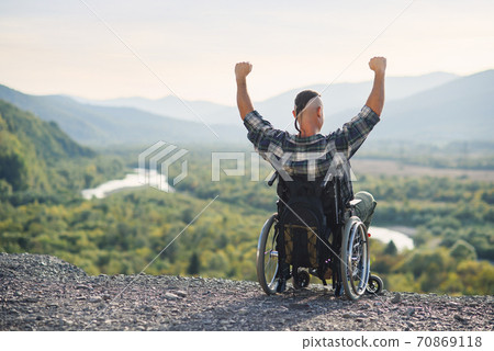 Disabled guy sitting in wheelchair with hands up on mountain in surrounded beautiful landscapes 70869118