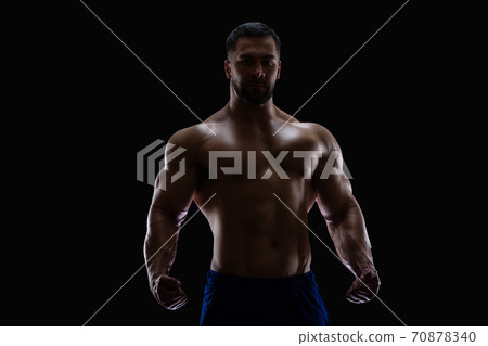 Portrait of a bodybuilder standing isolated on black background in a shadow with clenched fists to show off his muscles Portrait of a bodybuilder standing isolated on black background in a shadow with clenched fists to show off his muscles 70878340