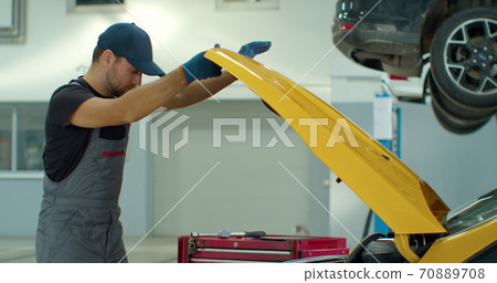 Professional car mechanic is working on a vehicle in a service. Happy repairman is closing the hood. Man Looks at a Camera and Smiles. 70889708