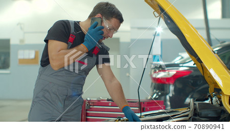 Mechanic talking on a mobile phone with the car owner while repairing a car in a garage. 70890941