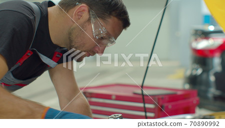 Close up footage of professional mechanic working on a vehicle in a car service. Engine specialist fixing the motor. Repairman is wearing gloves and using a ratchet. Close up footage of professional mechanic working on a vehicle in a car service. Engine specialist fixing the motor. Repairman is wearing gloves and using a ratchet. 70890992
