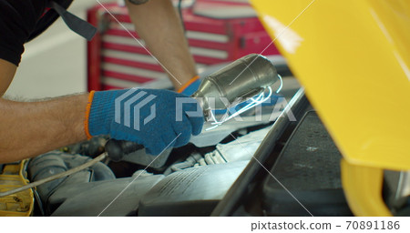 Repairman in safety glasses is working on an usual car maintenance. Professional mechanic is working on a car in a car service. He hangs a lamp and works with a ratchet. 70891186