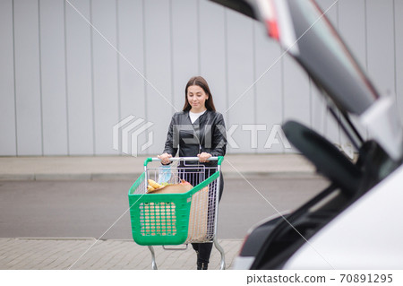 Young attractive woman walking with trolley groceries from a supermarket to car. Young attractive woman walking with trolley groceries from a supermarket to car. 70891295