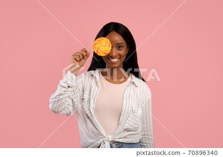 Smiling African American woman in casual clothes holding bright lollipop on pink studio background Smiling African American woman in casual clothes holding bright lollipop on pink studio background 70896340