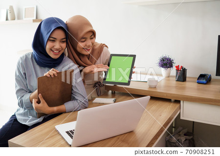 attractive cheerful young muslim business woman working on laptop tablet and smiling while sitting at her desk modern office with her friend 70908251