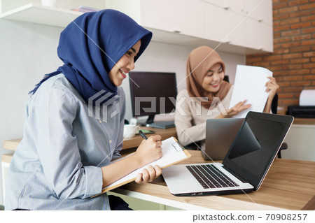 attractive cheerful young muslim business woman working on laptop and smiling while sitting at her desk modern office with her friend 70908257