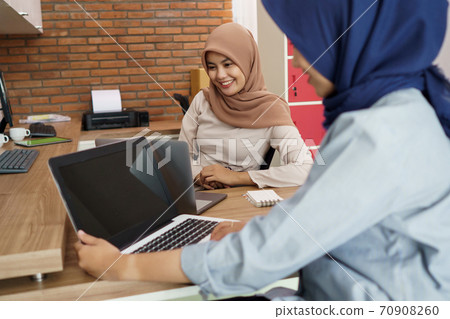 attractive cheerful young muslim business woman working on laptop and smiling while sitting at her desk modern office with her friend 70908260