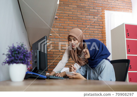attractive cheerful young muslim business woman working on computer and smiling while sitting at her desk modern office with her friend 70908297