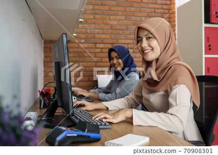 attractive cheerful young muslim business woman working on computer and smiling while sitting at her desk modern office with her friend 70908301