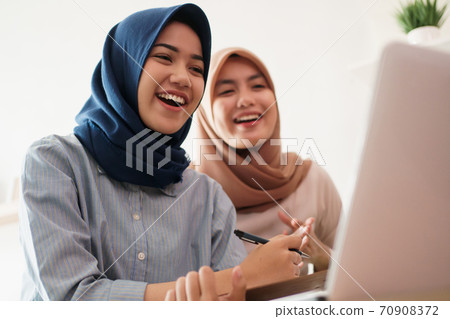 attractive cheerful young muslim business woman working on laptop and smiling while sitting at her desk modern office with her friend 70908372