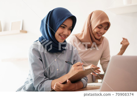 attractive cheerful young muslim business woman working on laptop and smiling while sitting at her desk modern office with her friend 70908374