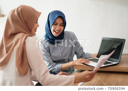 attractive cheerful young muslim business woman working on laptop and smiling while sitting at her desk modern office with her friend 70908394