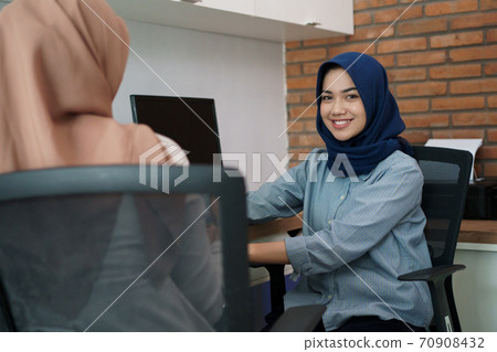 attractive cheerful young muslim business woman smiling while sitting at her desk modern office with her friend 70908432