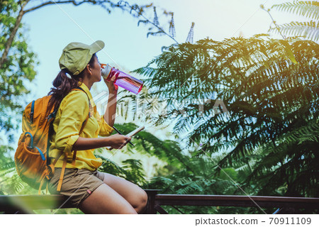 The young woman travels recording and studying the nature of the forest. She is sitting, relaxing and drinking water. The young woman travels recording and studying the nature of the forest. She is sitting, relaxing and drinking water. 70911109