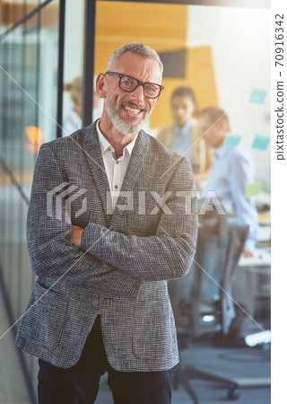 Vertical shot of a cheerful mature businessman in classic wear keeping arms crossed and smiling at camera while standing in the modern office Vertical shot of a cheerful mature businessman in classic wear keeping arms crossed and smiling at camera while standing in the modern office 70916342