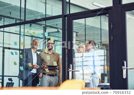 Group of creative coworkers standing in the modern office and looking at colorful sticky notes on a glass wall, discussing project Group of creative coworkers standing in the modern office and looking at colorful sticky notes on a glass wall, discussing project 70916352