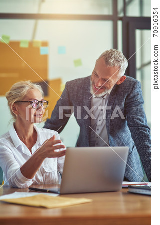 Two cheerful mature colleagues looking at laptop screen and discussing project results while working together in the modern office Two cheerful mature colleagues looking at laptop screen and discussing project results while working together in the modern office 70916354