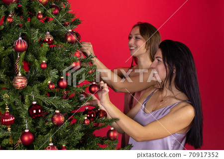 Happy lesbian couple joyfully decorate Christmas tree together on red backdrop Happy lesbian couple joyfully decorate Christmas tree together on red backdrop 70917352