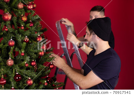 Young Gay Male Couple decorating Christmas Tree Together using ladder Young Gay Male Couple decorating Christmas Tree Together using ladder 70917541