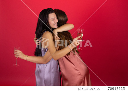 Two young lesbian women in elegant dresses hug holding champagne, red background Two young lesbian women in elegant dresses hug holding champagne, red background 70917615