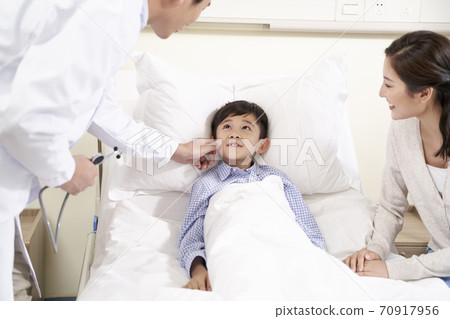 asian child lying in bed in hospital ward accompanied by mother talking to doctor 70917956