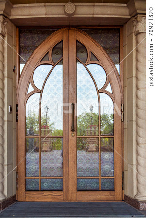 The front door on the facade of the building is wooden brown frame with glass inserts with a pattern, the double door in retro style. The front door on the facade of the building is wooden brown frame with glass inserts with a pattern, the double door in retro style. 70921820