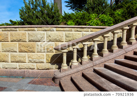 Baroque staircase with marble steps and stone balustrades under the railing on the background of a wall of stone tiles and plants in the background. 70921936