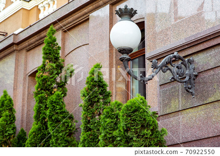 Forged iron street lamp with a white glass ceiling mounted on the marble wall of the facade of the building. Forged iron street lamp with a white glass ceiling mounted on the marble wall of the facade of the building. 70922550