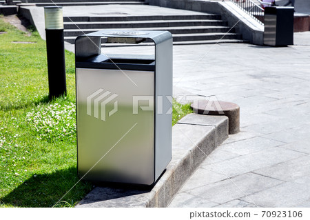A square outdoor trash can of metal is installed on the curb between a lawn with green grass and a pedestrian pavement of stone tiles against the background of the stairs. A square outdoor trash can of metal is installed on the curb between a lawn with green grass and a pedestrian pavement of stone tiles against the background of the stairs. 70923106