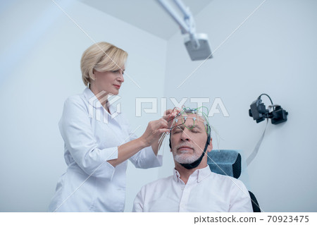 Female neurologist placing electrode cap on gray-haired male patient head Female neurologist placing electrode cap on gray-haired male patient head 70923475