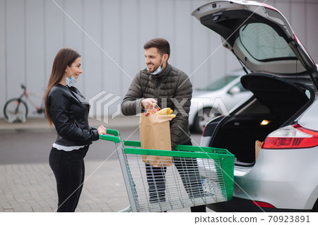 Young couple in masks loading bags in trunk after supermarket shopping during quarantine. Man with her wife in outdoor parking. Eco package with fresh food 70923891