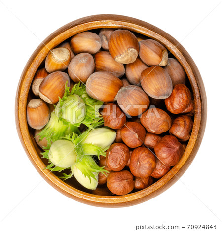 Ripe and unripe hazelnuts in a wooden bowl. Green, unripe hazelnuts in husk with shelled and unshelled hazelnuts. Seeds of Corylus avellana. Close-up from above, over white, isolated macro food photo. 70924843