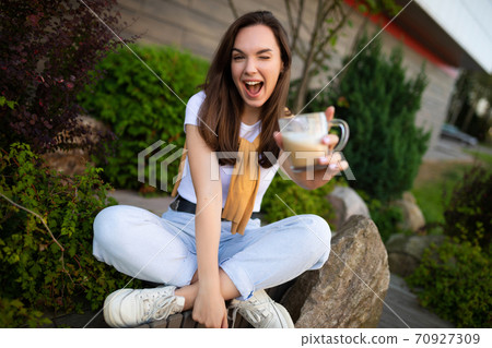 happy young woman sitting in a beautiful park and drinking spicy latte 70927309