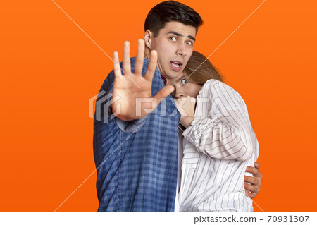 Do not come closer. Frightened female hiding at man chest, guy is terrified, makes stop sign with his hand Do not come closer. Frightened female hiding at man chest, guy is terrified, makes stop sign with his hand 70931307