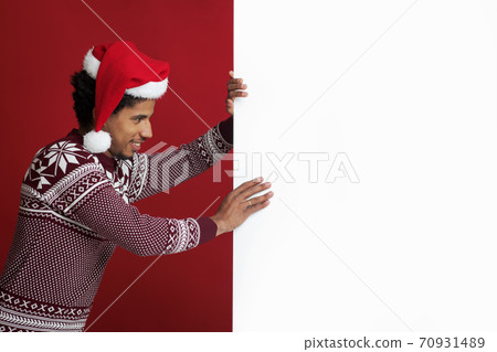 Millennial african american man in Santa Claus hat and sweater pushes white board with empty space 70931489