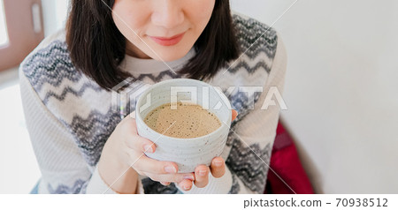 Young adult Asian woman wearing a knitted sweater enjoy drinking a hot drink in the morning in a white room background. One happy smiling girl relaxing indoors. With copy space 70938512