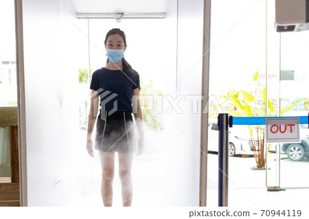 Asian child girl wear face mask,automatic disinfection tunnel is spraying on body for disinfecting at gate check point for COVID-19,measures to prevent,process of cleaning,new normal under Coronavirus 70944119