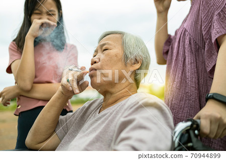 Asian senior woman holding a cigarette smoking,smokers elderly smoking near people in family,daughter, granddaughter closing nose,pollution from cigarettes smoke,dangerous to health,addiction,life 70944899