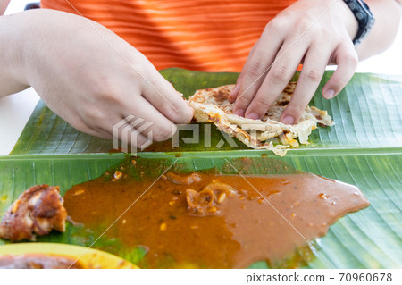 Person enjoying eating roti canai served with curry on banana leaf using hand 70960678