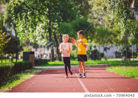 Couple of kids boy and girl doing cardio workout, jogging in park on jogging track red. Cute twins runing together. Run children, young athletes. Teen brother and sister running along path outdoors Couple of kids boy and girl doing cardio workout, jogging in park on jogging track red. Cute twins runing together. Run children, young athletes. Teen brother and sister running along path outdoors 70964048