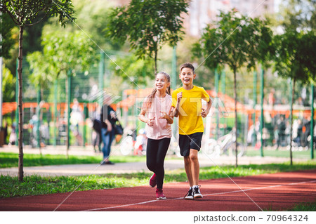 Couple of kids boy and girl doing cardio workout, jogging in park on jogging track red. Cute twins runing together. Run children, young athletes. Teen brother and sister running along path outdoors Couple of kids boy and girl doing cardio workout, jogging in park on jogging track red. Cute twins runing together. Run children, young athletes. Teen brother and sister running along path outdoors 70964324