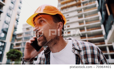 Angry male architect in orange hard hat talks swears on phone at construction site. Builder in plaid shirt emotionally displeased negotiates by cell phone. Screaming stressed architectural designer Angry male architect in orange hard hat talks swears on phone at construction site. Builder in plaid shirt emotionally displeased negotiates by cell phone. Screaming stressed architectural designer 70964401