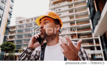 Angry male architect in orange hard hat talks swears on phone at construction site. Builder in plaid shirt emotionally displeased negotiates by cell phone. Screaming stressed architectural designer 70964413
