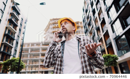 Angry male architect in orange hard hat talks swears on phone at construction site. Builder in plaid shirt emotionally displeased negotiates by cell phone. Screaming stressed architectural designer Angry male architect in orange hard hat talks swears on phone at construction site. Builder in plaid shirt emotionally displeased negotiates by cell phone. Screaming stressed architectural designer 70964446