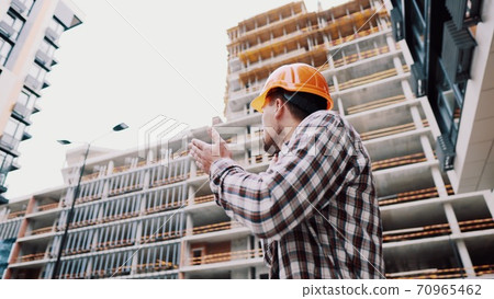 Portrait of young handyman making call while standing at construction area. Engineer talking on the phone on a construction site. Builder in helmet control according to plan by mobile phone Portrait of young handyman making call while standing at construction area. Engineer talking on the phone on a construction site. Builder in helmet control according to plan by mobile phone 70965462
