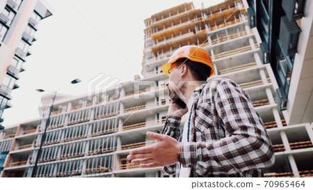 Caucasian male construction worker in orange hard hat and plaid shirt talking on phone at construction site. Architecture theme. Male profession. Foreman controls construction process by smartphone Caucasian male construction worker in orange hard hat and plaid shirt talking on phone at construction site. Architecture theme. Male profession. Foreman controls construction process by smartphone 70965464