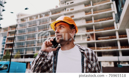 Portrait of young handyman making call while standing at construction area. Engineer talking on the phone on a construction site. Builder in helmet control according to plan by mobile phone Portrait of young handyman making call while standing at construction area. Engineer talking on the phone on a construction site. Builder in helmet control according to plan by mobile phone 70965518