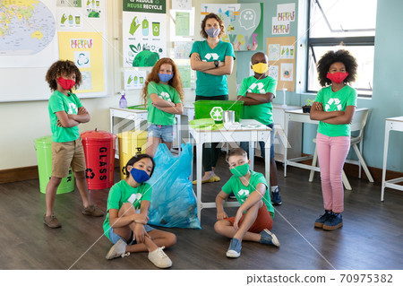 Female teacher and students wearing face masks with recycling container on table in class 70975382